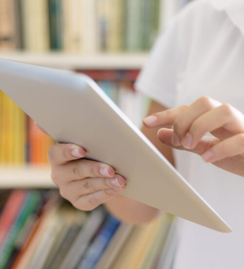 Female hands, making use of tablet computer Female hands, holding tablet computer, bookshelves at background - library or college class or hospital interior.